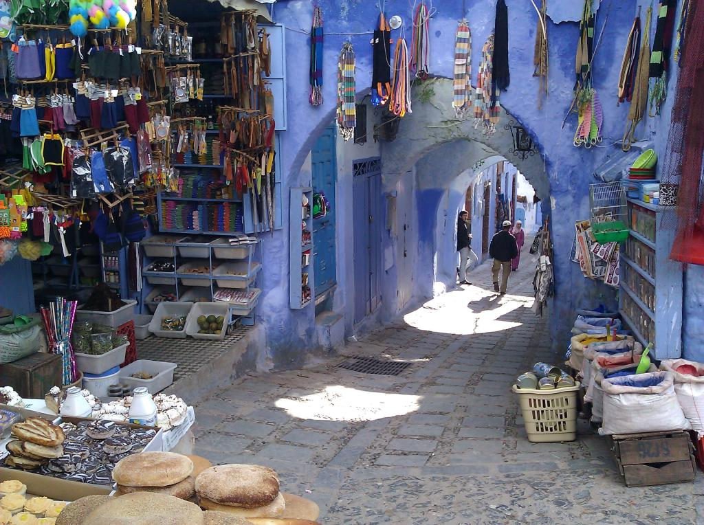 Ruelle bleue étroite avec des étals colorés et des tissus suspendus, des passants, sur des pavés ensoleillés : une scène vivante et colorée.