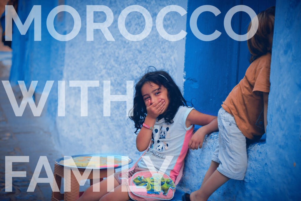 Girl smiles, covers mouth, sitting by blue wall with another child nearby. Text âMorocco with Familyâ overlays. Playful mood.