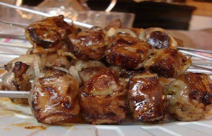 Grilled, Moroccan boulfaf, glazed meat skewers on a white plate; rich, golden-brown hues in a kitchen setting. Appears appetizing and savory.
