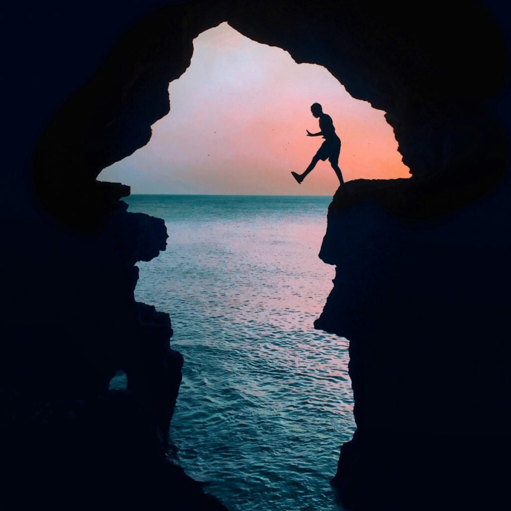 Silhouette of a person jumping between rock formations at hercules caves in tangier morocco at sunset by the sea. Sky is pink and orange, creating a serene backdrop.