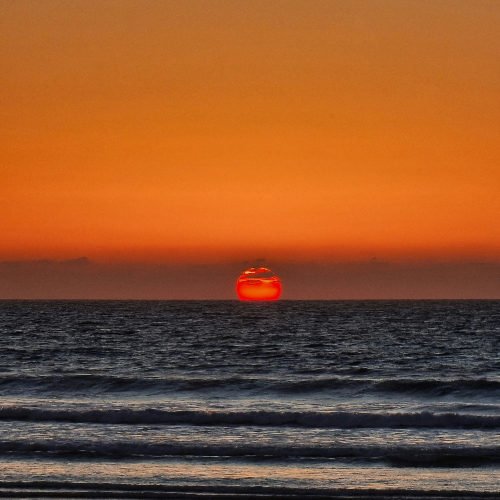 Sunset over ocean with vivid orange sky, red sun at horizon, and dark, rippling waves creating a calm, serene atmosphere.