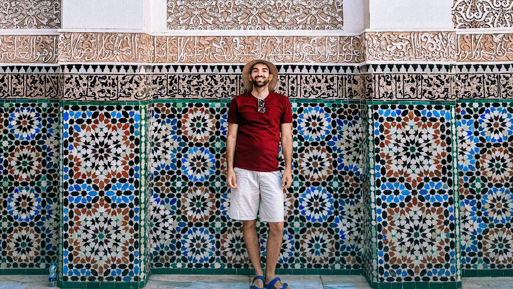 Man in a hat and red shirt stands smiling against a colorful, intricate tiled wall with ornate Arabic calligraphy above in morocco. Bright, relaxed setting.