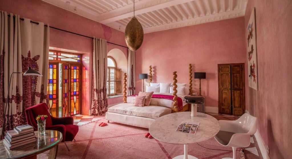 Pink bedroom with a plush bed, stained glass doors, and a marble table. A red chair holds flowers, and there’s warm, elegant decor.