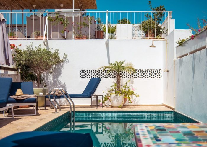 A serene poolside with blue loungers, potted plants, and white walls under a clear sky. A colorful table edge is visible in the foreground.
