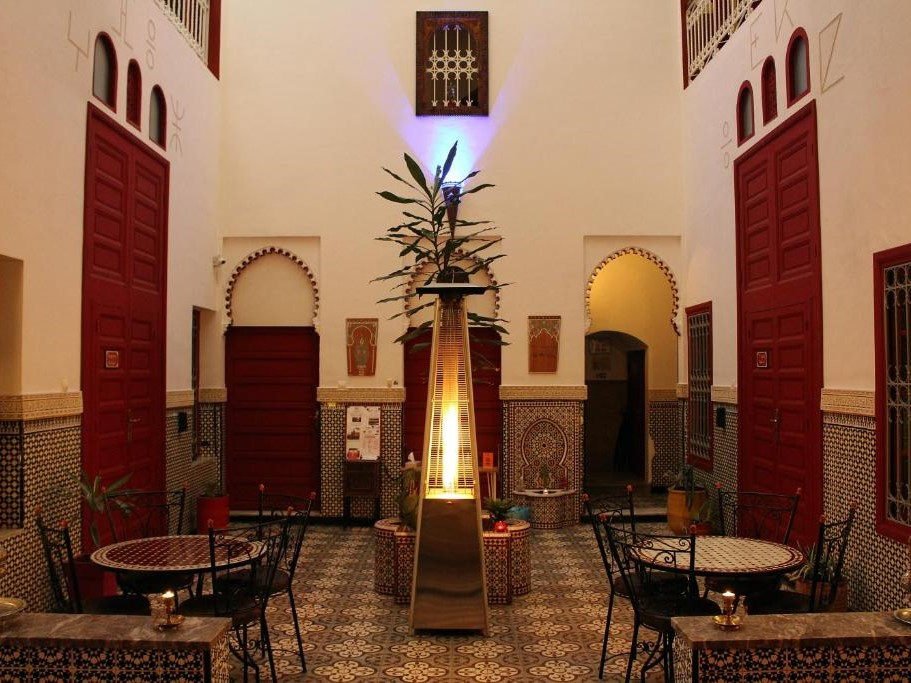 Elegant courtyard with round tables, chairs, and a central tall heater. Red doors and ornate patterns on walls create a warm ambiance.