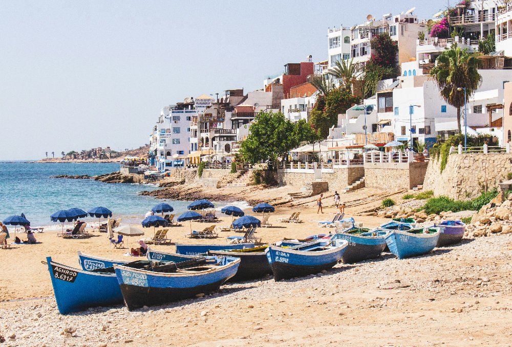Fishing boats on a sandy beach, blue umbrellas scattered, and people walking. White buildings line the coast under a clear, sunny sky in Taghazout Morocco.