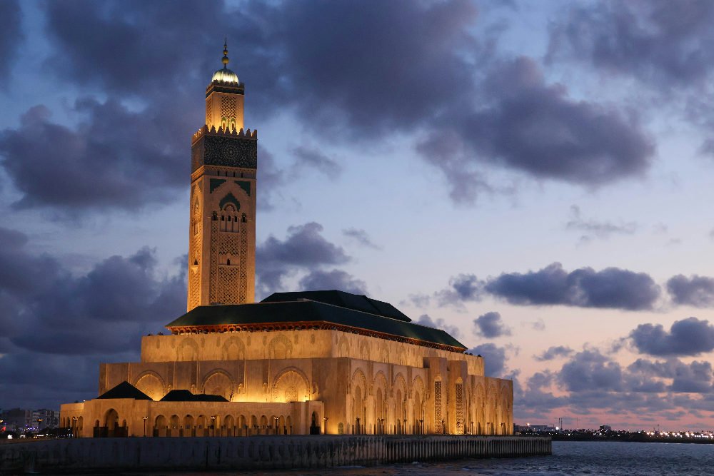 Illuminated mosque with tall minaret at sunset, surrounded by water. Cloudy sky casts a peaceful mood. No visible text.