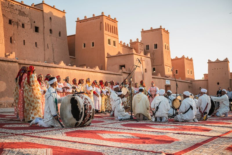 A group in colorful attire performs ahwach music and dance on patterned rugs in Morocco, set against ancient tan-colored buildings under a clear sky.