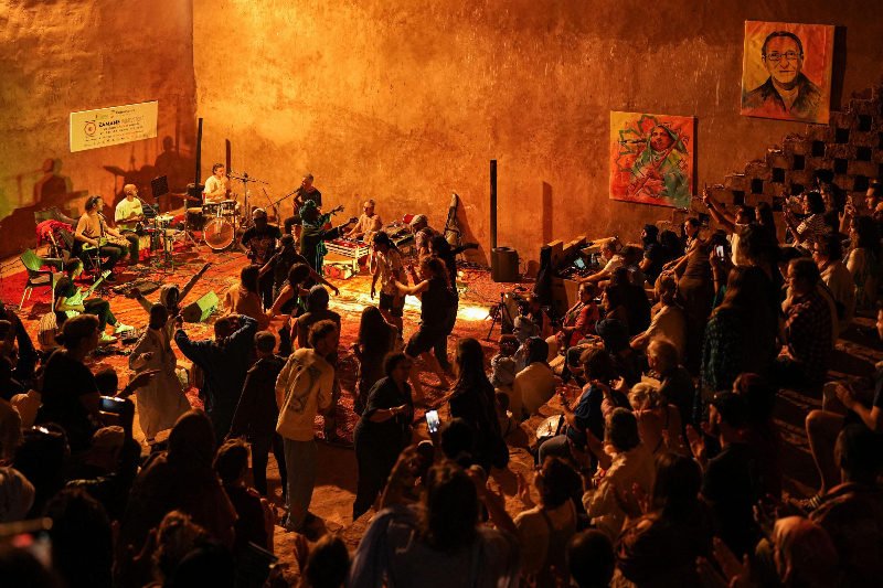 Concert in warm-lit hall, musicians on stage with guitars and drums in the moroccan sahara, crowd dancing and clapping, colorful artwork on walls. Energetic mood.