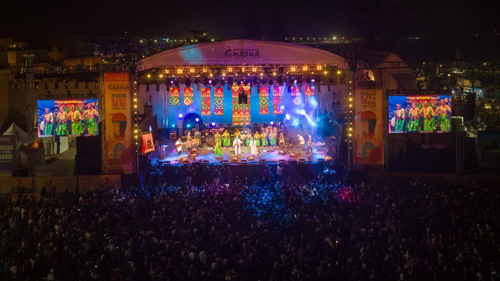 Night concert with musicians on stage, bright lights, colorful patterns above, and a large crowd below during essaouira gnaoua festival in morocco. Screens display performers. Festive mood.