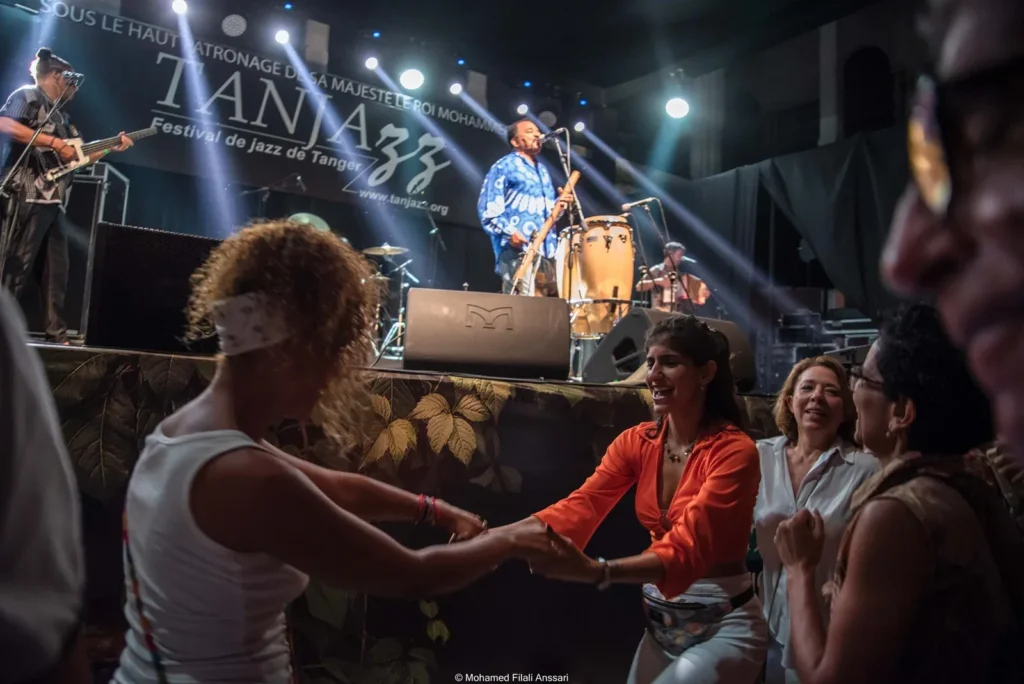 Crowd dancing joyfully in front of a band on stage at Tanjazz festival. Bright lights and vibrant colors create an energetic atmosphere.